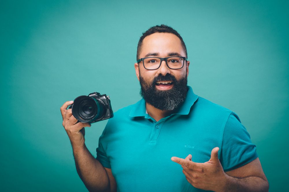 Der Rostocker Fotograf Martin Börner steht gutgelaunt im Fotostudio in Evershagen. Mit Brille und Bart hält er eine Nikon-Kamera in der Hand. Er trägt ein türkisfarbenes Poloshirt und steht vor einem türkisfarbenen Hintergrund.